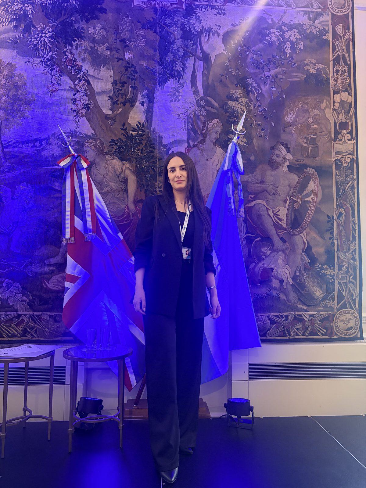 Vanina Sciarrone de pie en un escenario, frente a un tapiz y las banderas del Reino Unido y Argentina